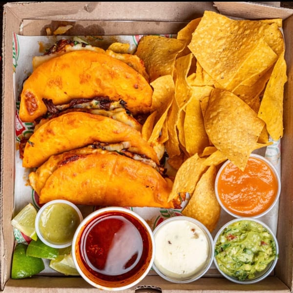 Quesa Loco birria tacos, tortilla chips, guacamole, and various sauces in a takeout box.