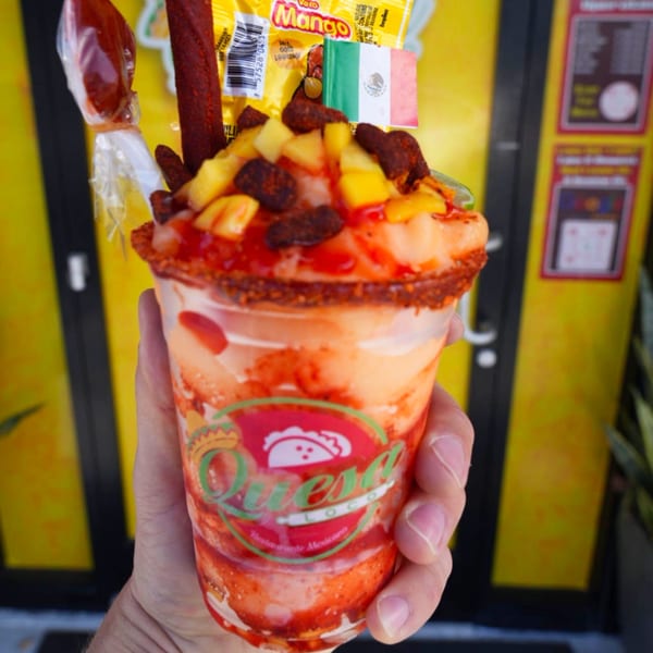 Hand holding a Quesa Loco mango chamoyada with chili candy and a small Mexican flag.