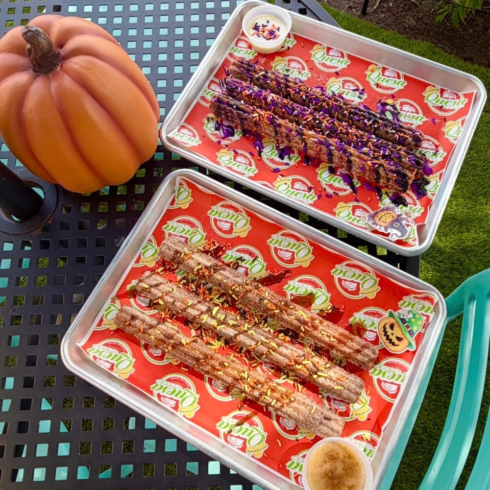 Two trays of chocolate-covered churros with colorful sprinkles and dipping sauces beside a pumpkin on an outdoor patio table.