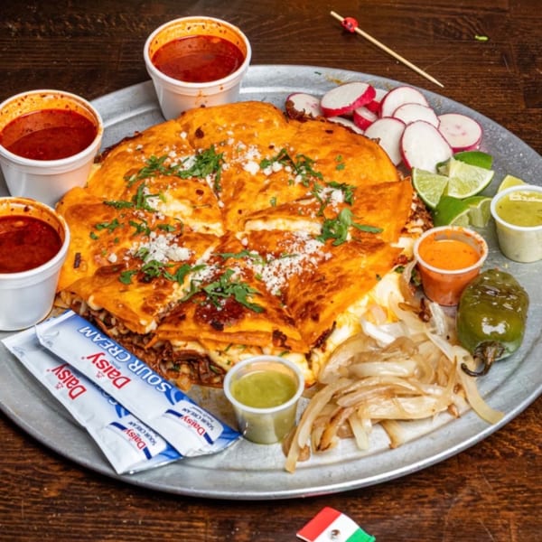 A large Quesa Loco quesadilla with sides of consommé, radishes, limes, grilled onions, and sauces.