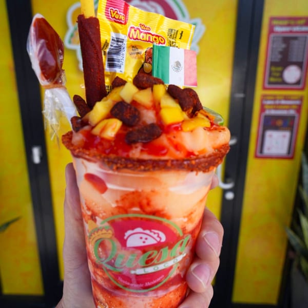 Hand holding a Quesa Loco mango chamoyada with candy, a Mexican flag, and a yellow background.