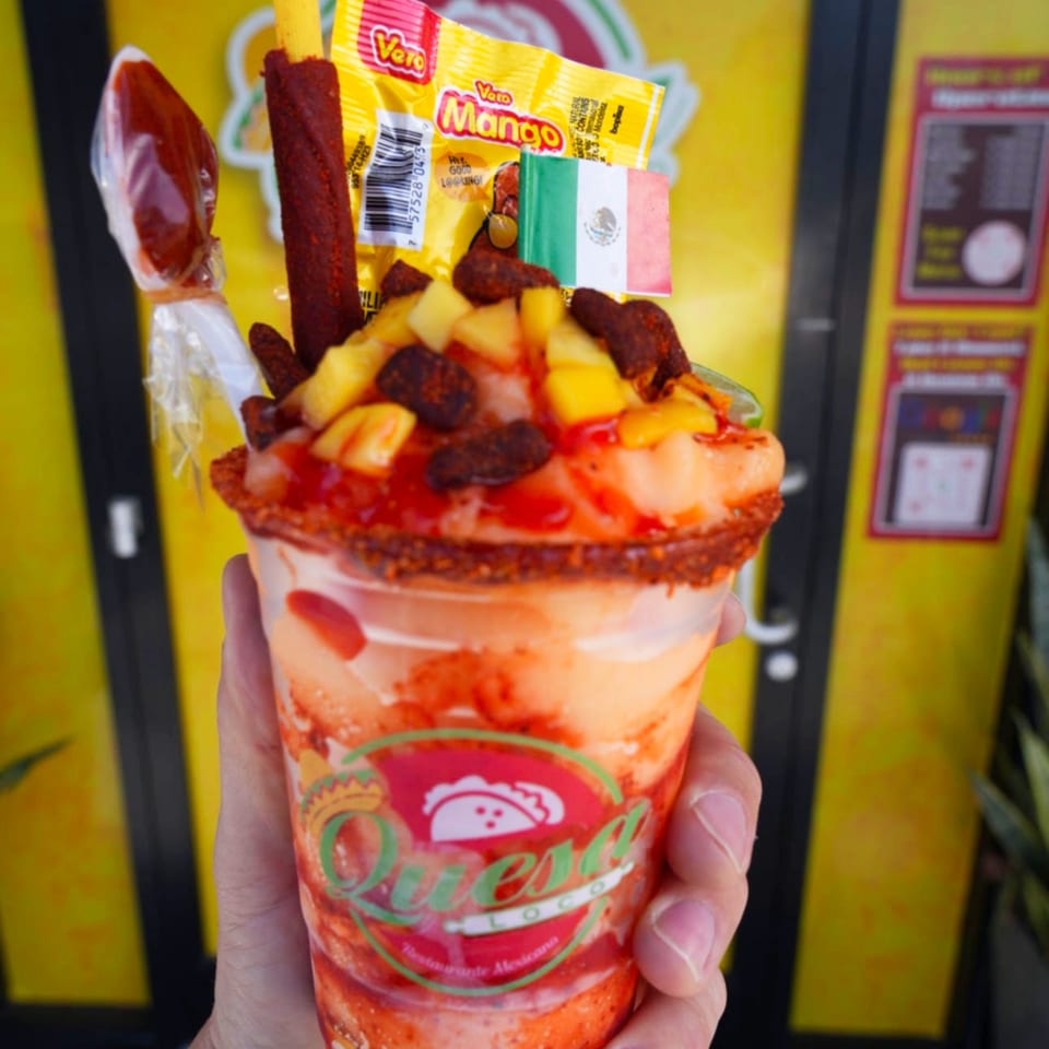 Hand holding a Quesa Loco mango chamoyada with candy, a Mexican flag, and a yellow background.