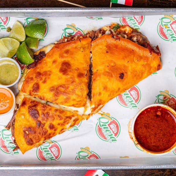 Two large, cheesy Quesa Loco birria quesadillas with limes, green and orange sauces, and a side of consommé.