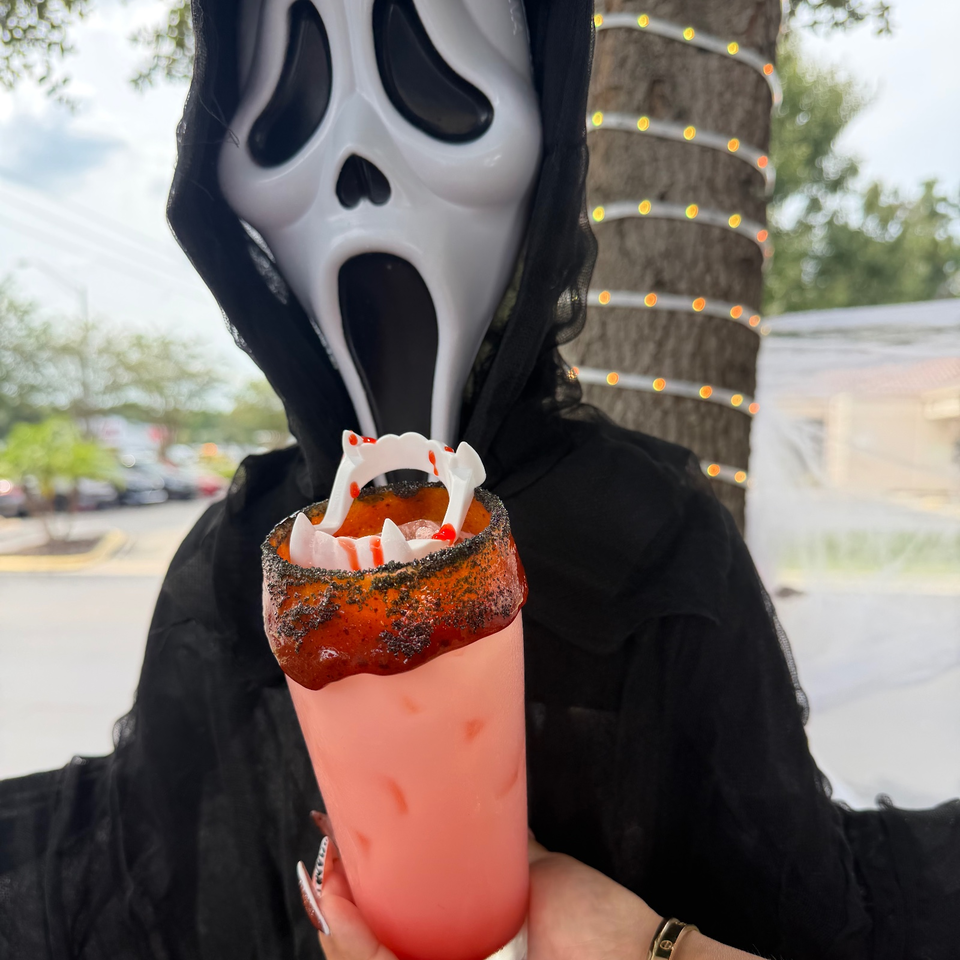 Person wearing a Ghostface mask holds a red cocktail with a black rim, vampire fangs, and fake blood garnish outdoors.