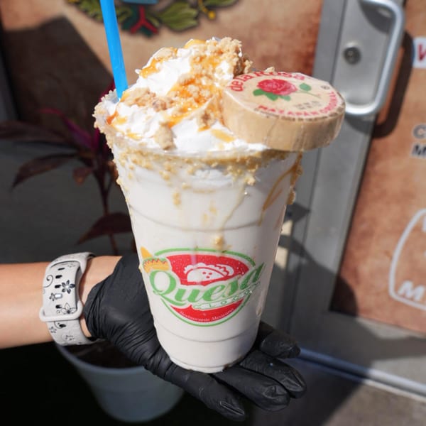 Hand in black glove holding a Quesa Loco milkshake topped with whipped cream, caramel, and a De La Rosa mazapan.