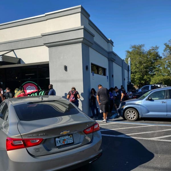 People queue outside Quesa Loco, a Mexican restaurant, with cars parked in the foreground.