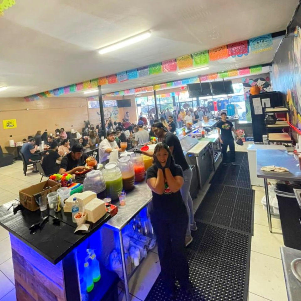 A bustling Quesa Loco restaurant interior with customers dining and staff behind a counter with colorful drinks.