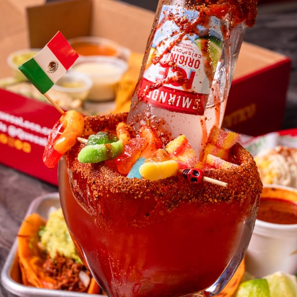 A Quesa Loco michelada with a Smirnoff Ice bottle, candy, and a Mexican flag, next to tacos.