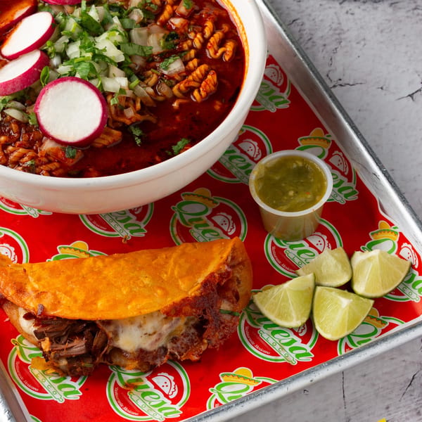 Quesa Loco birria ramen with radishes and onions, a birria taco, salsa, and lime wedges.