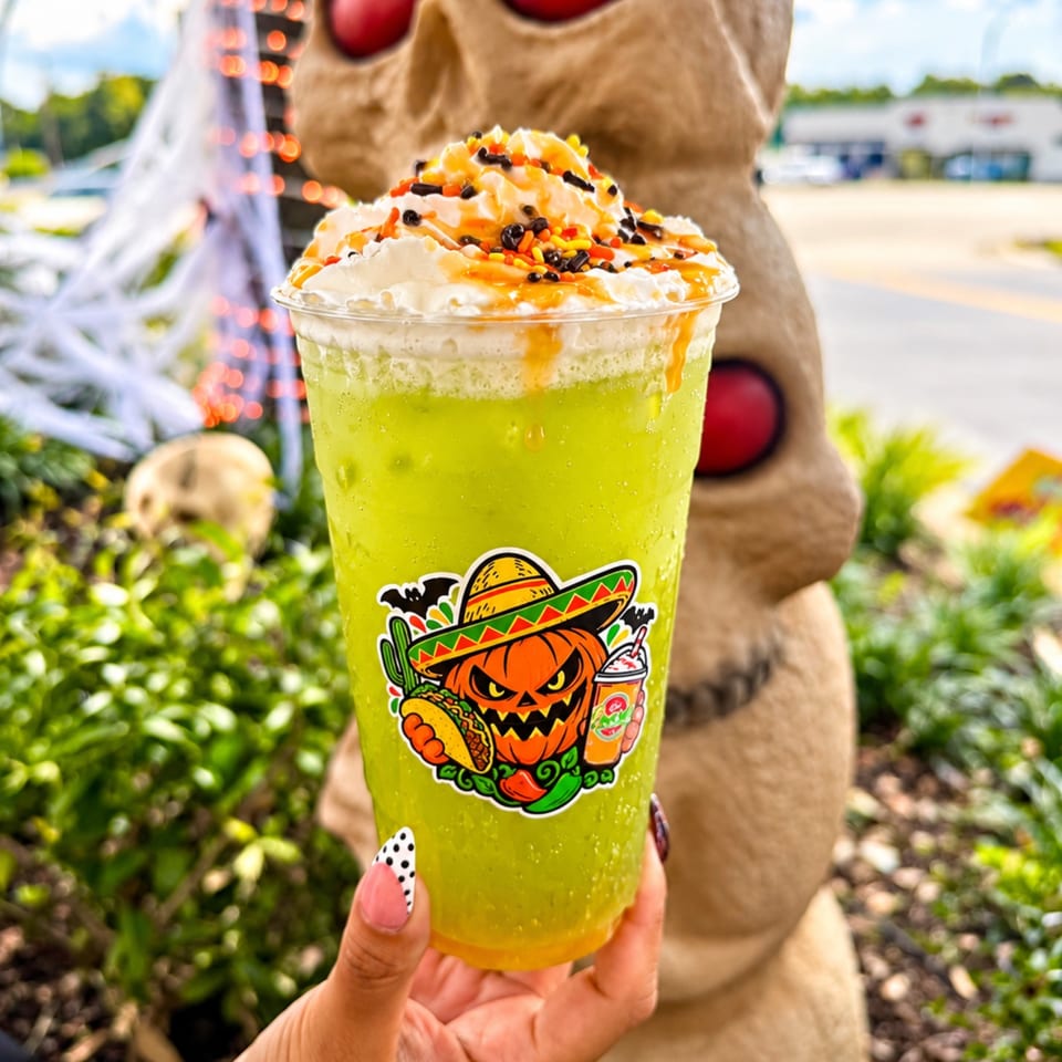 Bright green frozen drink topped with whipped cream, orange and black sprinkles, held outdoors beside a Halloween statue.