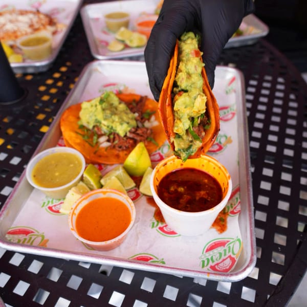 Hand in black glove holding a Quesa Loco taco filled with guacamole over a tray of food.