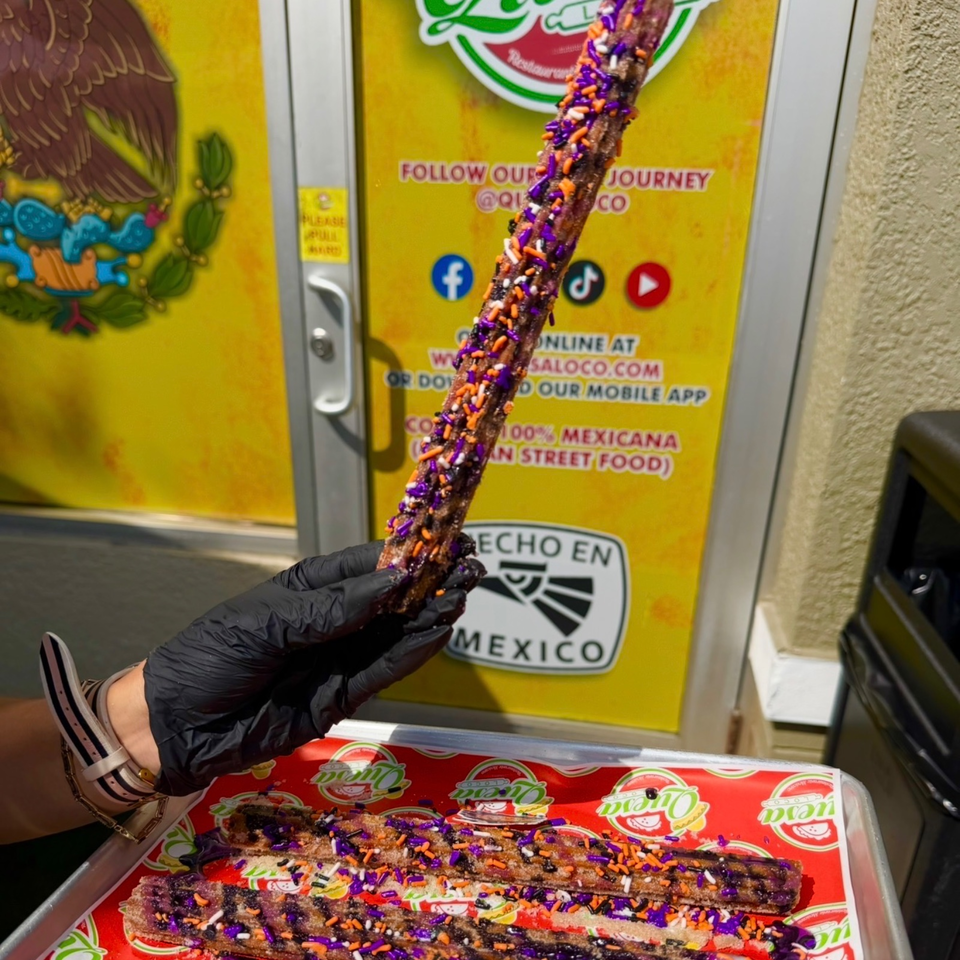 Gloved hand holds a chocolate-covered churro topped with purple, orange, and white sprinkles above a tray of decorated churros.