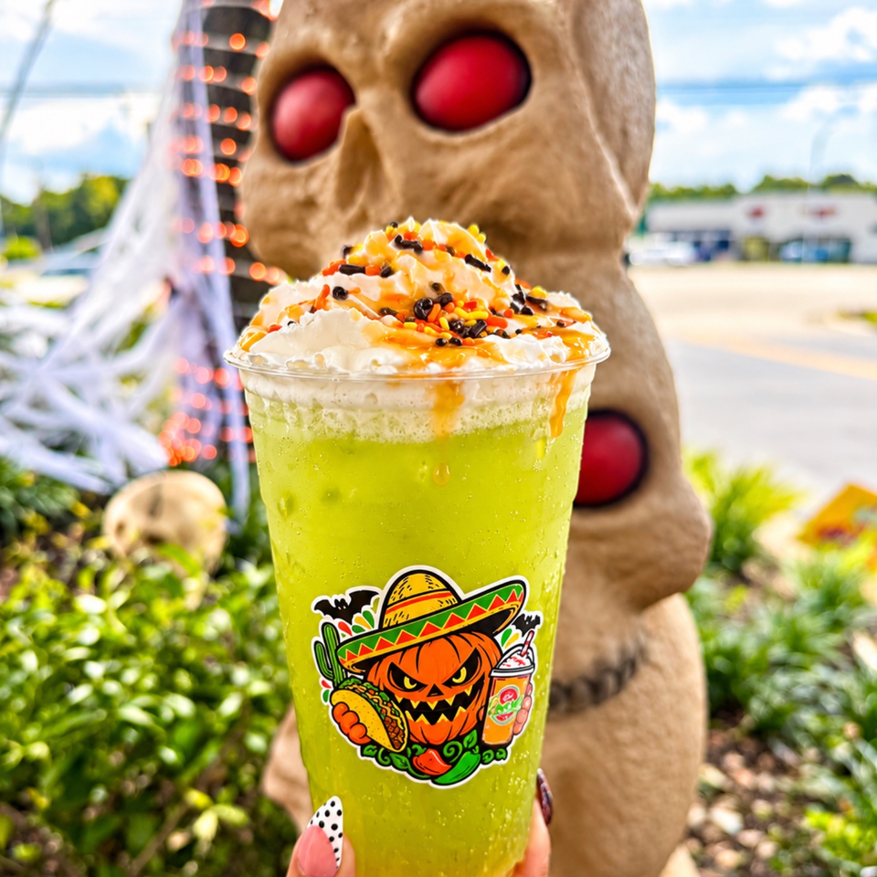 Bright green blended drink topped with whipped cream, orange drizzle, and colorful sprinkles, held before a large outdoor skull statue.