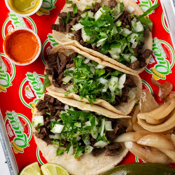 Three Quesa Loco carne asada tacos with cilantro and onions, served with sauces and limes.