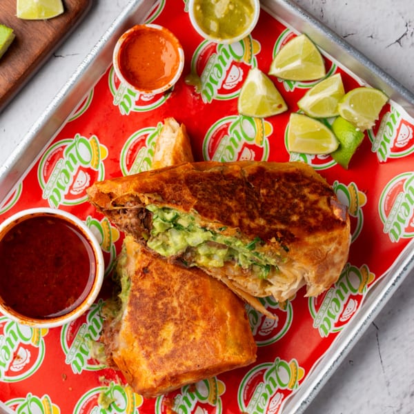 A Quesa Loco burrito, cut in half, revealing meat and guacamole, with sauces and lime wedges.