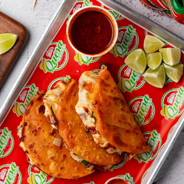 Three Quesa Loco birria tacos with consome and lime wedges on a branded red tray.