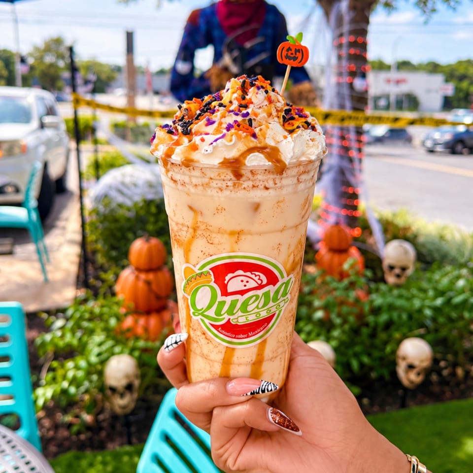 Queso-branded Halloween milkshake topped with whipped cream, caramel, and colorful sprinkles, held outdoors beside spooky decorations.
