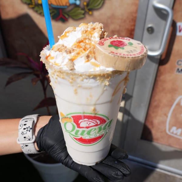A hand in a black glove holds a Quesa Loco milkshake topped with whipped cream, caramel, and a peanut candy.