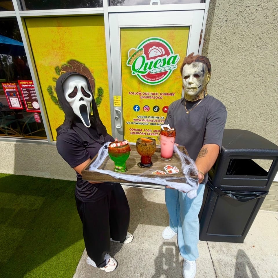Two costumed people stand outside Quesa Loco holding a tray of colorful drinks wrapped in decorative cobwebs.