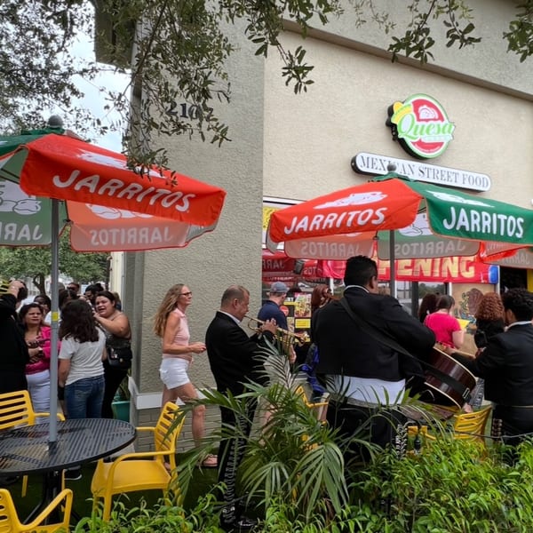 Mariachi band playing outside Quesa Loco, a Mexican street food restaurant, with patrons and Jarritos umbrellas.