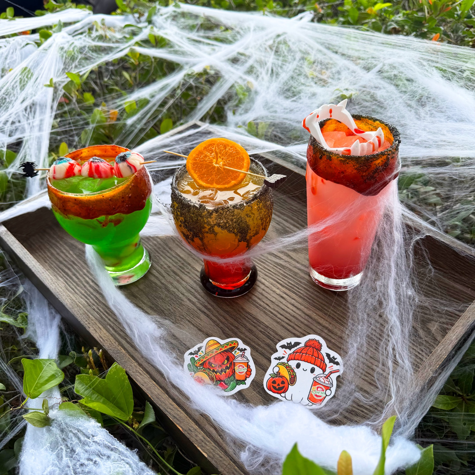 Three colorful Halloween cocktails sit on a wooden table draped in cobwebs, garnished with fruit, candy, and spooky decorations.