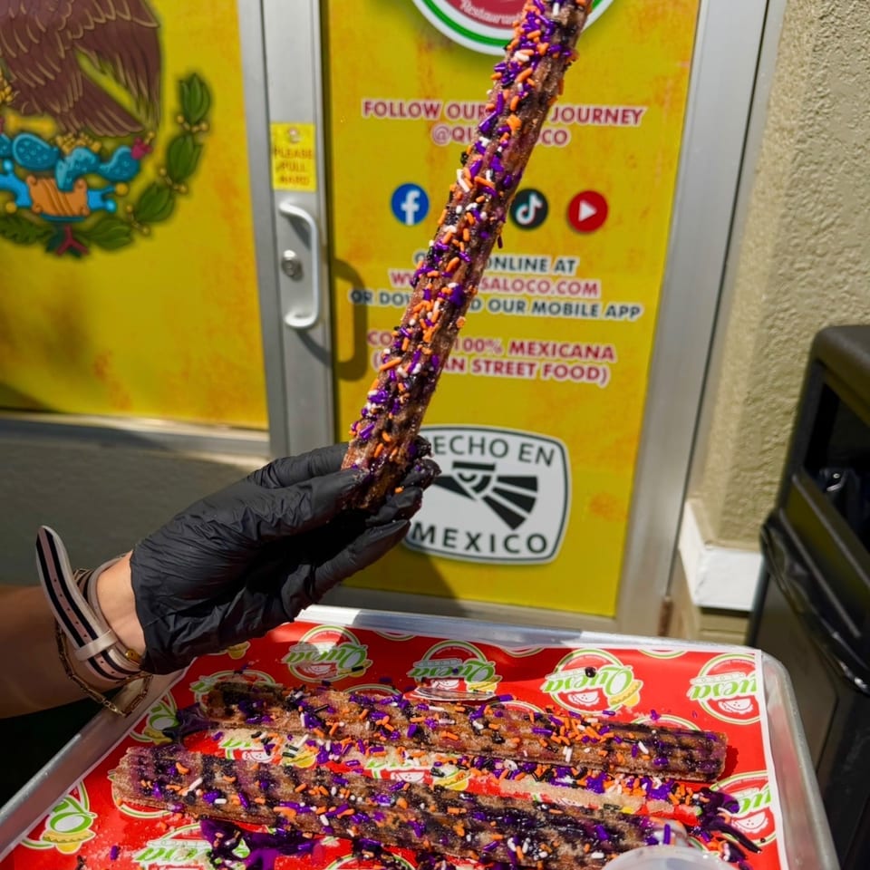 Gloved hand holds a chocolate-coated churro covered in purple, orange, white, and black sprinkles above a decorated tray.