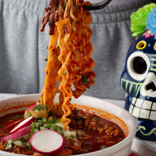 Fork lifting birria ramen noodles from a bowl with radishes, onions, and cilantro, next to a sugar skull.