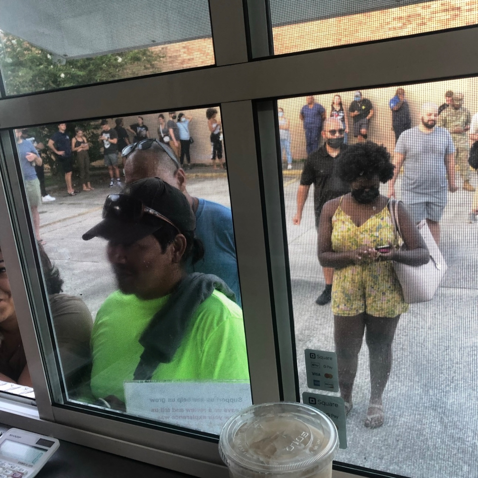 Customers wait in line outside Quesa Loco, viewed through a screened window with a drink and calculator.