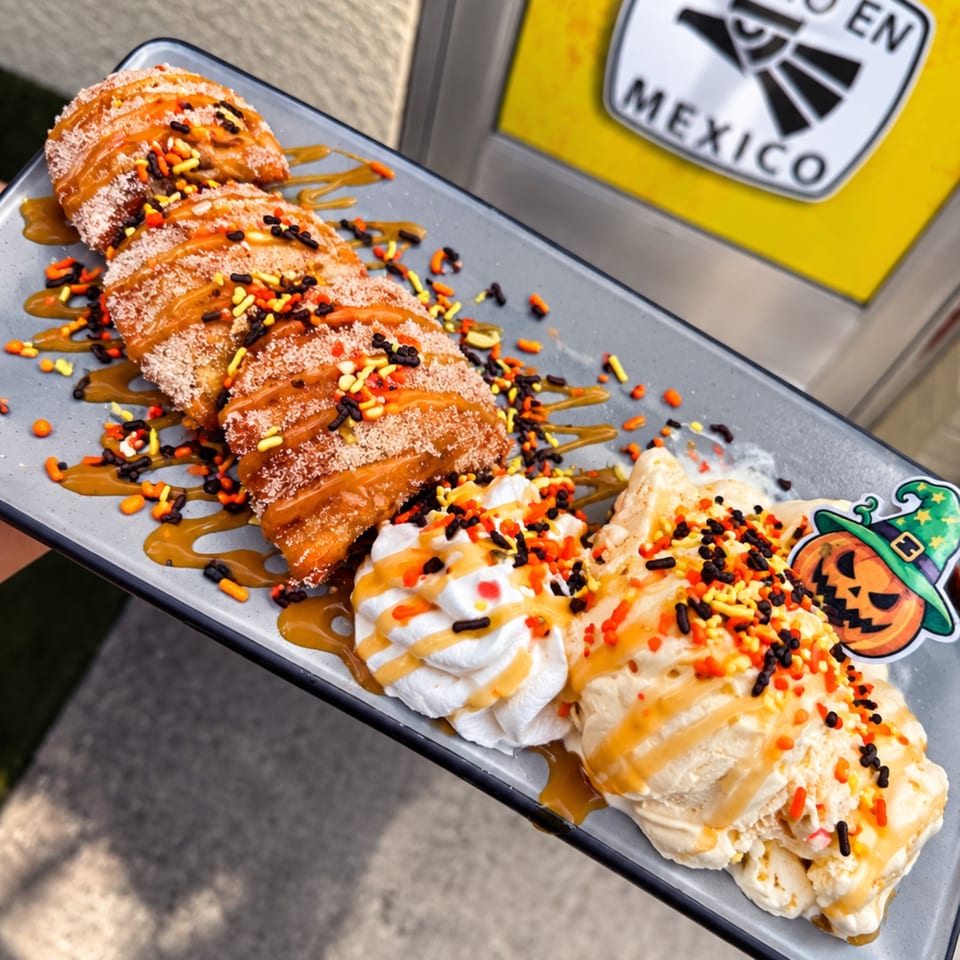 Sliced churro-style pastries drizzled with caramel, whipped cream, and orange, yellow, and black sprinkles on a platter.