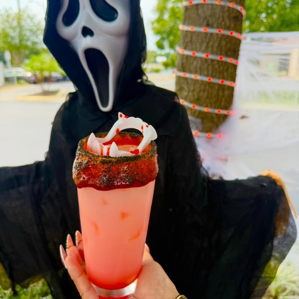 Pink cocktail with a spicy rim and gummy worms held before a person in a ghost mask and black costume.
