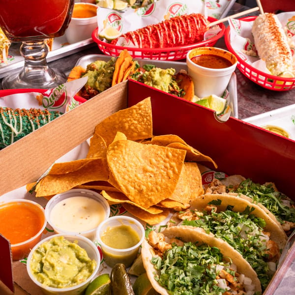 A Quesa Loco takeout box filled with tacos, chips, guacamole, and various sauces, alongside other Mexican dishes.